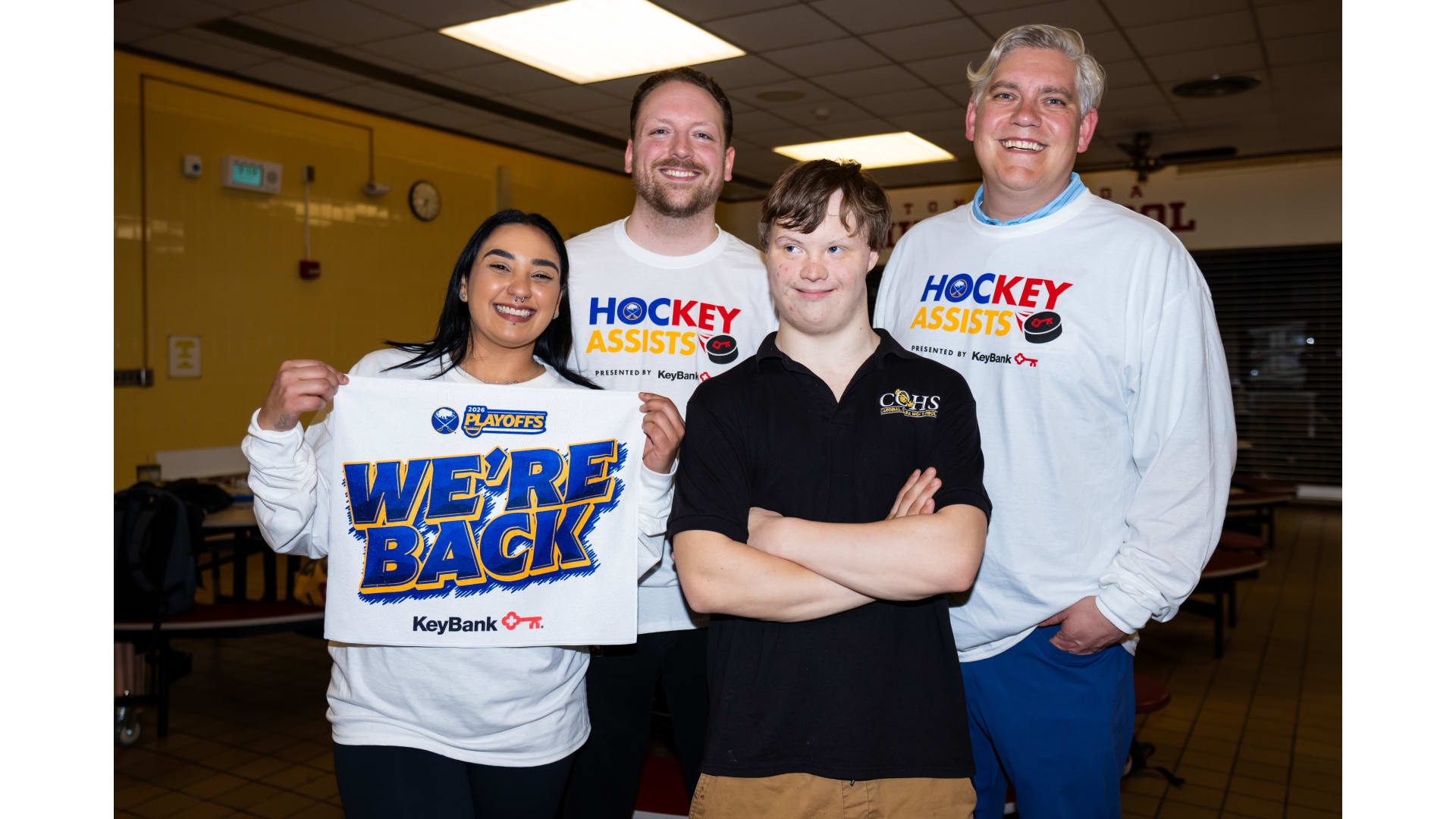 Four people standing together, one holding up a "we're back" banner