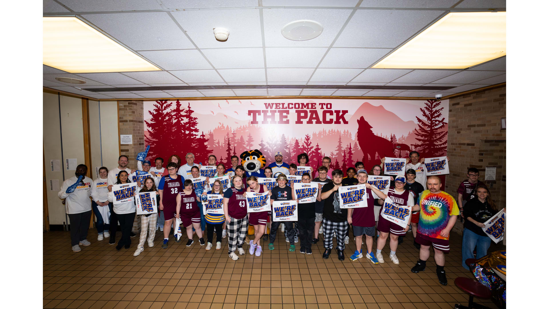 A larger group posing with "we're back" banners in front of a mural that says "Welcome to THE PACK"